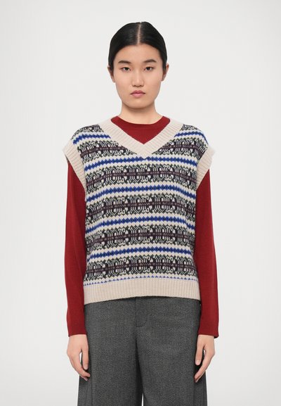 Gilet en tricot avec un décolleté en V, présentant des rayures à motifs multicolores en bleu, noir et crème. Porté par-dessus une chemise à manches longues rouge.