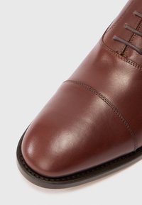 Zapato de vestir de cuero marrón con costura en la punta y suela oscura, mostrado en vista cercana sobre un fondo blanco.