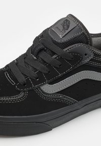 Scarpe da skate in suede nero con accenti grigi, dotate di un colletto imbottito, lacci piatti e perforazioni per la ventilazione. Suola in gomma con grip testurizzato.