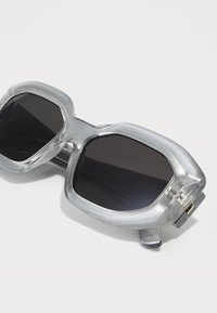 Lunettes de soleil surdimensionnées en argent avec une finition brillante, des verres rectangulaires et une monture angulaire proéminente. Présente des verres teintés en noir et des accents métalliques.