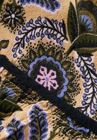 Tissu floral présentant des motifs complexes en bleu, vert et rose sur un fond beige, avec des bords noirs. Tissage texturé visible.