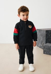 Black zip-up sweatshirt with colorful red and green stripes, featuring a tiger logo. Paired with black pants and white sneakers.