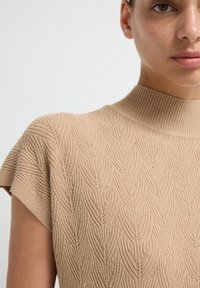 Top de puntobeige con un escote alto, que presenta un patrón de hojas texturizado y mangas tipo cap sleeve, resaltando un tejido ligero y texturizado.