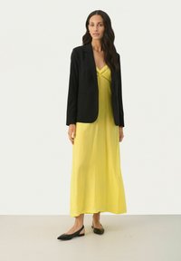 Schwarzer Blazer über einem gelben Maxikleid mit figurbetontem Oberteil, Spitzenakzenten und einem fließenden Rock. Kombiniert mit schwarzen flachen Schuhen.