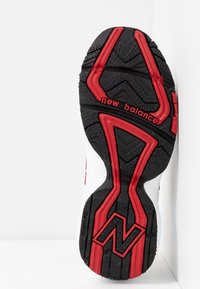 Semelle de chaussure New Balance noire et rouge avec une bande de roulement texturée ainsi que du texte et un logo de marque en bas, orientés vers le haut sur fond blanc.