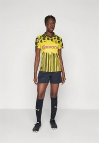 Dres Borussia Dortmund v rumeni barvi z črnimi poudarki in črtastim vzorcem, kombiniran s črnimi hlačami in nogavicami, z blagovno znamko Puma.