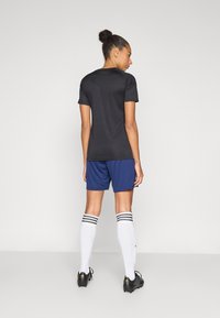 Γυναίκα με τα μαλλιά σε κότσο φοράει μαύρο μπλουζάκι με κοντά μανίκια, μπλε σορτς, λευκές κάλτσες μέχρι το γόνατο με μαύρες ρίγες και μαύρα ποδοσφαιρικά παπούτσια, στέκεται με την πλάτη προς τα εμπρός.