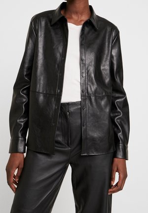 Osoba ubrana w czarną koszulę z imitacji skóry na guziki, na białej bluzce, w połączeniu z dopasowanymi czarnymi skórzanymi spodniami.