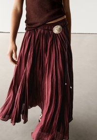 Jupe longue bordeaux froissée avec une ceinture ajustée, dotée d'une boucle circulaire argentée et d'un tissu texturé, tombant fluidement jusqu'au sol.