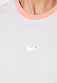 Camiseta de color gris claro con una textura suave. Presenta un cuello redondo de color rosa pálido y un logotipo de Nike plateado reflectante en el pecho.