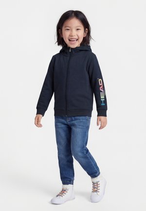 Nasmejan otrok v črni jopa z zadrgo in logotipom "HEAD" na rokavu, modrih kavbojkah in belih visokih supergah z pisanimi vezalkami.