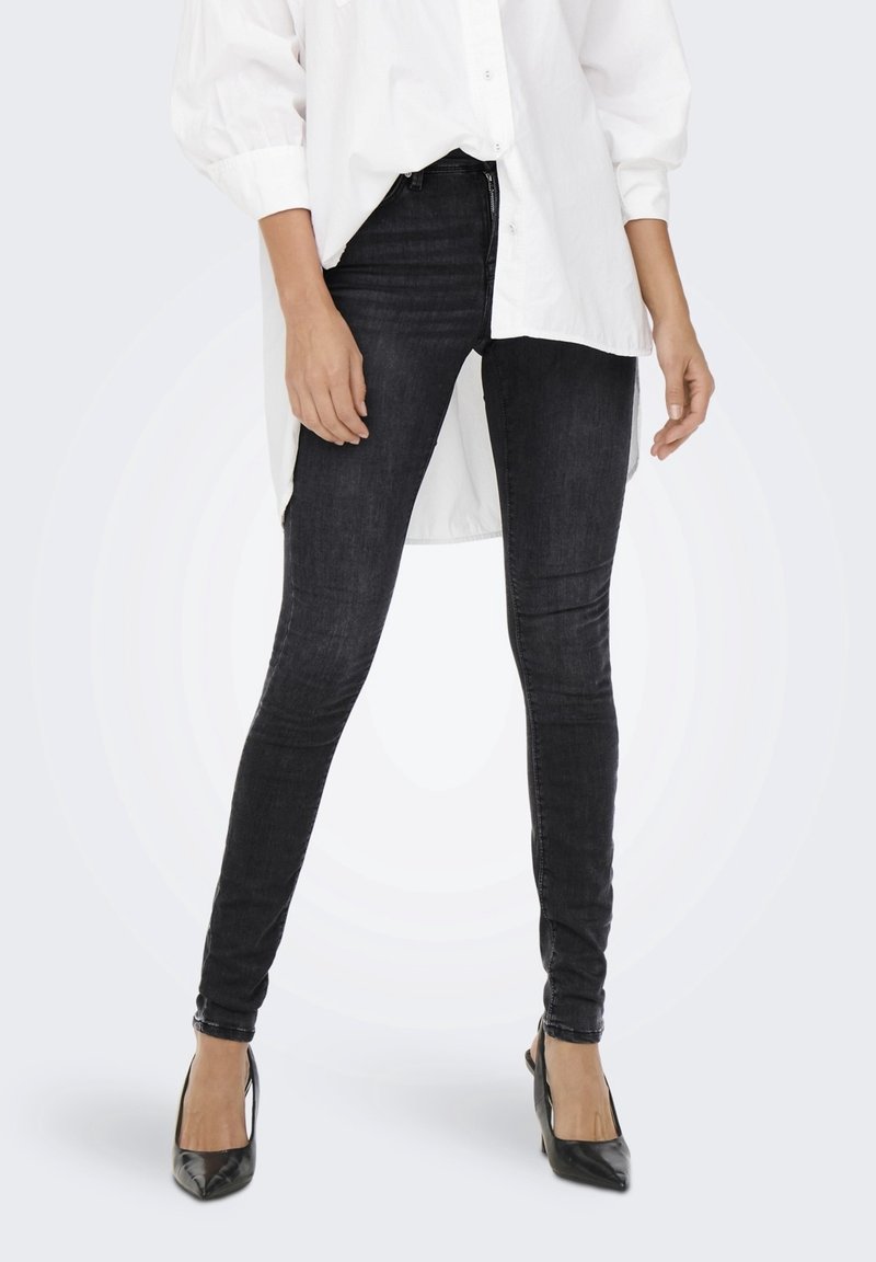 Jeans skinny negros de denim, con un ajuste ceñido en las piernas, complementados con una camisa blanca de gran tamaño y tacones negros.