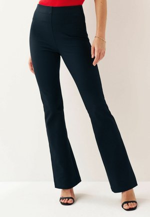 Femme portant un pantalon noir taille haute évasé, des sandales noires à bouts ouverts et un haut rouge, debout sur un sol blanc devant un fond uni.