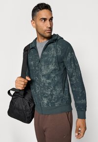 Tamnozelena zip-up dukserica s teksturiranim uzorcima, nosi se preko sive majice. Upareno s smešnim duksericama i crnom torbom s mrežastim detaljima.
