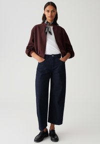 Giacca bomber marrone sopra una camicia bianca, abbinata a jeans larghi di jeans scuri e scarpe stringate nere. Accessoriata con una sciarpa a righe.