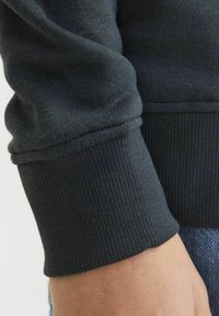 Mračni rukav od dukserice s rebrastom teksturom. Krupni plan pokazuje meki materijal i elastičnu završnicu, ističući detalje šivanja.