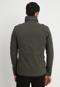 Oliven grøn jakke med høj krave, skulderepauleter og et struktureret stof. Har et tætsiddende design med sidelommer.