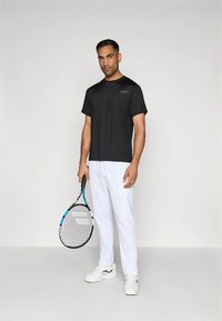 T-shirt atlética preta de manga curta com gola redonda, combinada com calças brancas e ténis brancos. A pessoa segura uma raquete de ténis.