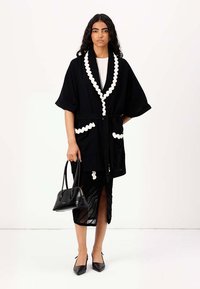 Cardigan noir avec des empiècements en dentelle blanche, manches courtes, ceinture à nouer et poches avant, associé à une jupe noire transparente et des escarpins pointus.
