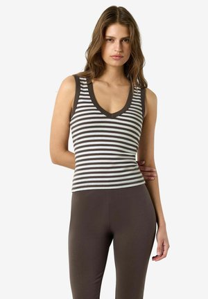 Femme portant un haut sans manches à rayures noires et blanches avec un col en V et un pantalon ajusté marron foncé uni sur un fond blanc uni.