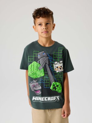 Băiat purtând un tricou verde închis cu tematică Minecraft, cu personaje din joc și grafice de blocuri, stând pe un fundal simplu și deschis la culoare.