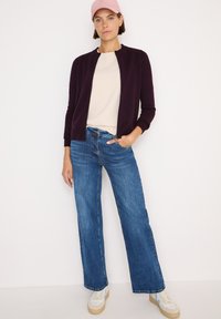 Cardigan a maglia bordeaux con collo rotondo, abbinato a una maglietta beige e jeans a gamba larga blu. Completato con sneakers bianche e un cappellino rosa.