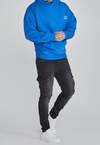 Felpa con cappuccio blu, dal taglio largo, abbinata a pantaloni cargo neri con tasche laterali e sneakers alte bianche.