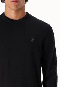 Maglione nero a maglia con scollatura a girocollo, polsini a coste e un piccolo logo ricamato sul petto. Tessuto morbido e design aderente.