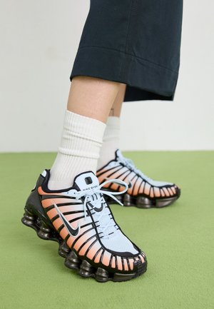 Stopy w białych skarpetkach i czarnych spodniach, w butach Nike Shox z białymi i pomarańczowymi detalami na zielonej podłodze.