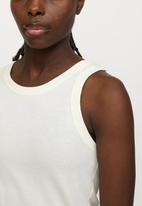 Weißes, geripptes Tanktop mit rundem Ausschnitt, kontrastierender cremefarbener Naht, figurbetonter Passform und einer weichen, glatten Textur.