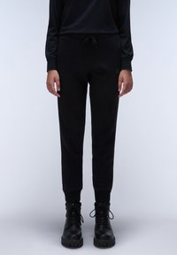 Zwarte jogpants van zachte stof met een elastische tailleband en drawstring. Aansluitende boorden en subtiele logo-detail aan de zijkant. Gepaard met zwarte laarzen.