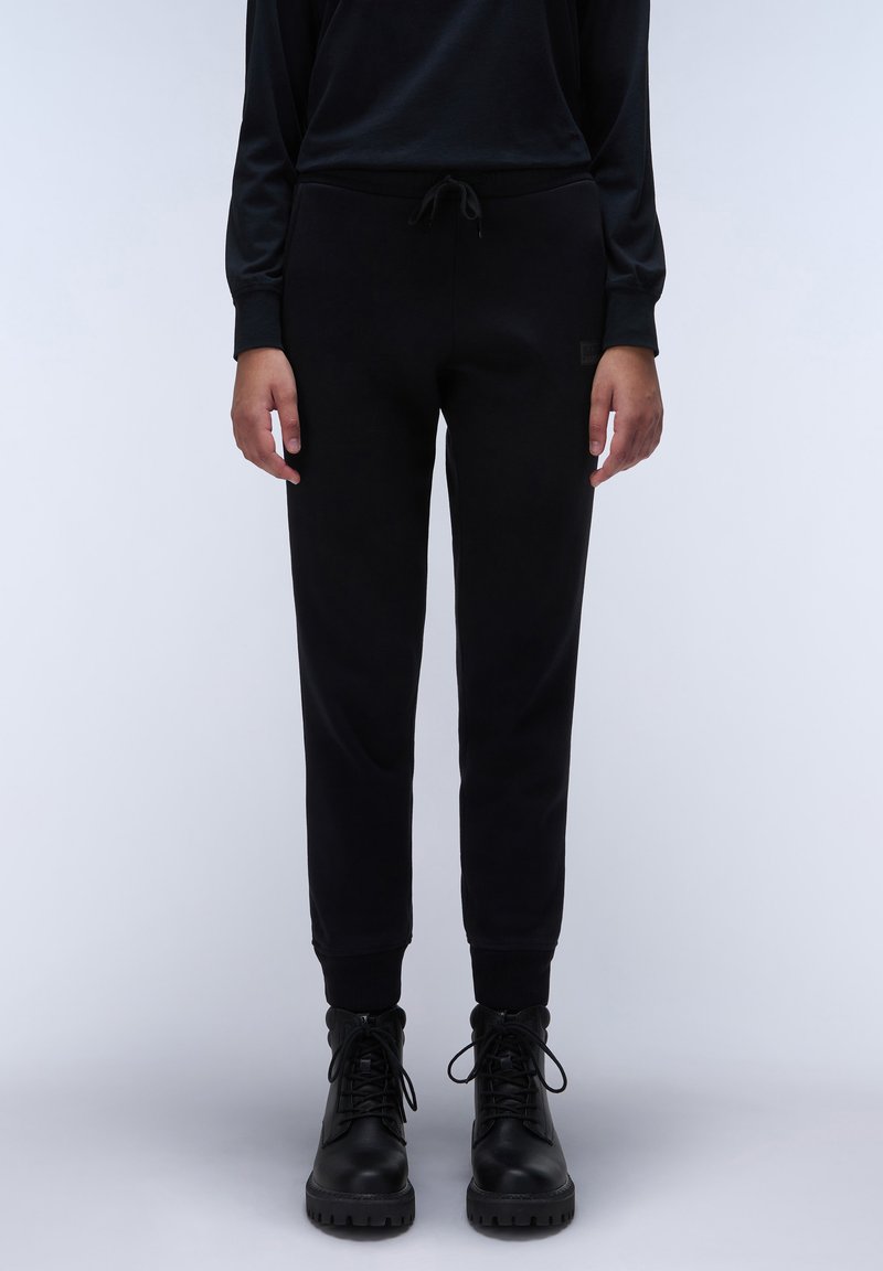 Zwarte jogpants van zachte stof met een elastische tailleband en drawstring. Aansluitende boorden en subtiele logo-detail aan de zijkant. Gepaard met zwarte laarzen.
