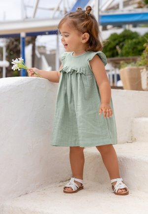 Robe en coton vert clair avec des manches volantes et un corsage froncé, associée à des sandales blanches à lanières. L'enfant tient de petites fleurs.
