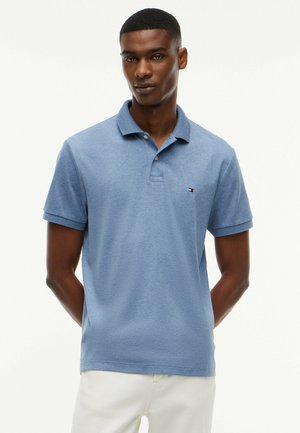 Junger Mann trägt ein hellblaues kurzärmeliges Polohemd mit kleinem Logo auf der Brust und weiße Hose vor einfachem Hintergrund.