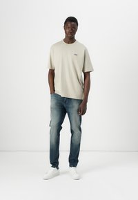Camiseta de manga corta gris claro con logo en el pecho, combinada con jeans azules desgastados y zapatillas blancas, de pie contra un fondo liso.