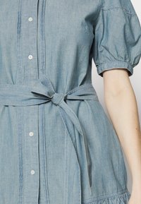 Džínové šaty v světle modré barvě s krátkými nabíranými rukávy, předním zapínáním na knoflíky a zavazovacím pasovým páskem. Obsahují nařasený volánový lem.