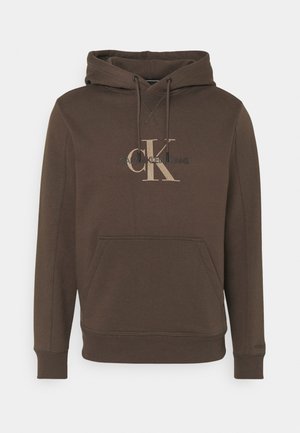 Mężczyzna w czarnej skórzanej kurtce na brązowej bluzie z kapturem Calvin Klein, czarnych szortach, czarnych wysokich trampkach i czarnych skarpetkach na jednolitym tle.