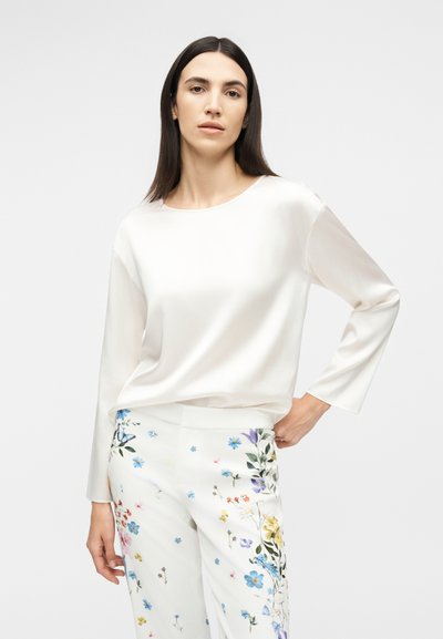 Γυναίκα με μακριά σκούρα μαλλιά, φορώντας λευκό μακρυμάνικο τοπ και λευκό παντελόνι με πολύχρωμα λουλουδάτα σχέδια, ποζάρει μπροστά σε απλό φόντο.