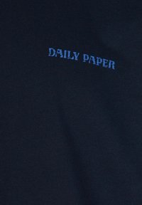 Maglietta blu navy in tessuto testurizzato, con il testo "DAILY PAPER" ricamato in un blu chiaro sbiadito sul lato superiore sinistro.