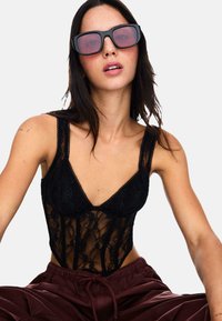 Schwarzer Spitzenbody mit zarten Blumenmustern, dünnen Trägern und einem tiefen Ausschnitt. Das Model trägt übergroße schwarze und rote Sonnenbrillen.