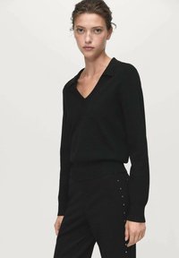 Crni pulover s v-neck izrezom i dugim rukavima, izrađen od mekog materijala; prilagođen u struku; nosi se s odgovarajućim hlačama s dekorativnim zakovicama.