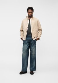 Homme debout les mains dans les poches, portant une veste beige, une chemise noire, un jean large bleu avec des détails de couture, et des chaussures noires, fond neutre.