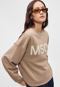 Καφέ φούτερ με υπερμεγέθη εφαρμογή, με κεντημένο λογότυπο "MSC" σε πιο ανοιχτό τόνο. Το μοντέλο φοράει γυαλιά ηλίου με λεοπαρντί σχέδιο.