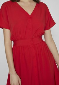 Czerwona sukienka wykonana z lekkiego materiału, z dekoltem w kształcie litery V, krótkimi rękawami i regulowanym pasem w talii. Posiada frontowe guziki i zwiewny dół.