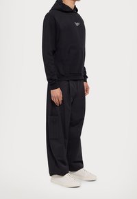 Czarna bluza z kapturem z przednią kieszenią, zestawiona z luźnymi czarnymi spodniami z boczną kieszenią oraz białymi trampkami.
