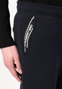 Navy blue sweatpants feature a zippered side pocket, contrasting white accents, and a branded logo along the pocket edge. Soft material texture.