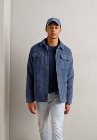 Veste en velours côtelé bleu avec deux poches poitrine, portée sur un pull sombre. Un jean bleu clair et une casquette bleue assortie complètent la tenue.