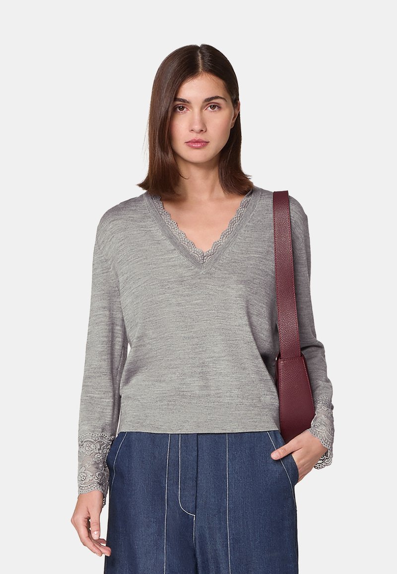 Grauer, gestrickter Pullover mit V-Ausschnitt, gewellten Details am Kragen und an den Ärmelbündchen, kombiniert mit weiten, dunkelblauen Jeanshosen und einer kastanienbraunen Tasche.
