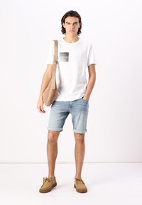 T-shirt blanche avec impression graphique, shorts en denim bleu clair et chaussures en suède beige. Porte un sac en toile beige à épaule.