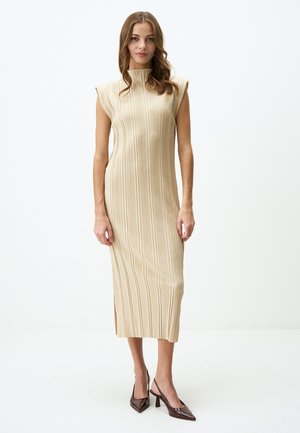 Mouwloze beige jurk met verticale gestreepte patroon, hoge halslijn, zijsplitten en een gladde textuur. Gecombineerd met donkerbruine hakken.